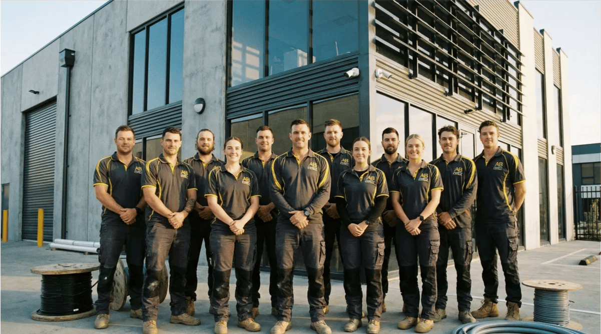 A group of twelve people wearing matching work uniforms stand in front of a modern industrial building, posing for a team photo that reflects their commitment to safety and quality electrical solutions.